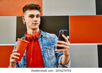 Young Caucasian Guy Drinking Soda Using Smartphone Leaning On The Wall