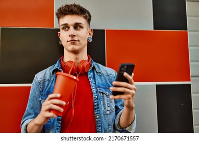 Young Caucasian Guy Drinking Soda Using Smartphone Leaning On The Wall