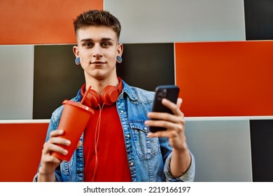 Young Caucasian Guy Drinking Soda Using Smartphone Leaning On The Wall