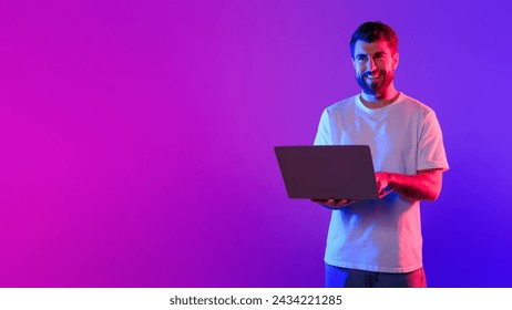 Young caucasian guy using laptop standing in studio with purple and blue neon light, showcasing concept of online work and gaming. Technology and freelance. Panorama, copy space - Powered by Shutterstock