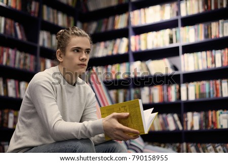 Young Caucasian guy wearing gray sweater spending day at bookstore searching for book on art, holding yellow one in his hands, having uncertain look. People, reading, leisure and knowledge concept