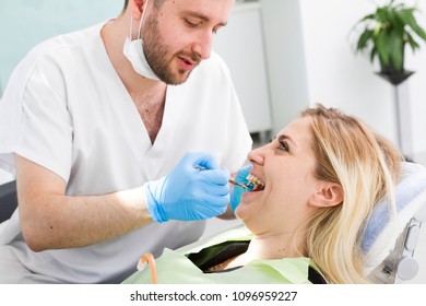 Young Caucasian Handsome Dentist in dental office talking with happy female blonde patient and preparing for treatment. Surgeon who specializes in dentistry, diagnosis, prevention, and treatment.
