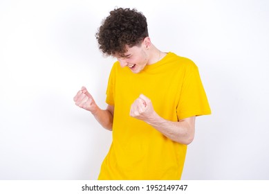 young caucasian handsome man with curly hair wearing yellow T-shirt against white studio background very happy and excited doing winner gesture with arms raised, smiling and screaming for success.