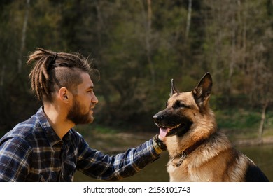 Young Caucasian hipster with dreadlocks and beard spends time with dog in park on sunny summer day. Concept of pet as family member. Owner and German Shepherd walk and look at each other devotedly.