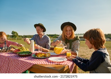 Young Caucasian Hipster Farm Family Having Lunch With Fresh Organic Products In Countryside Outdoors. Mother Pouring Juice For Son. Father And Daughter Eating Fruits. Modern Farm Lifestyle. Sunny Day