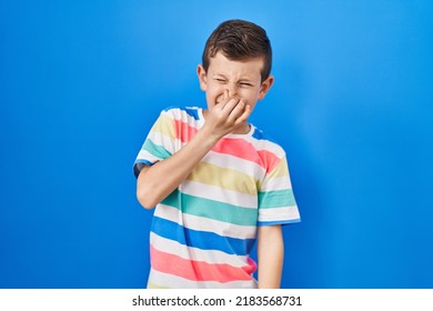 Young Caucasian Kid Standing Over Blue Background Smelling Something Stinky And Disgusting, Intolerable Smell, Holding Breath With Fingers On Nose. Bad Smell 