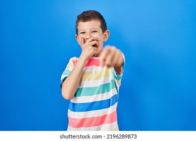 Young Caucasian Kid Standing Over Blue Background Laughing At You, Pointing Finger To The Camera With Hand Over Mouth, Shame Expression 