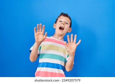 Young caucasian kid standing over blue background afraid and terrified with fear expression stop gesture with hands, shouting in shock. panic concept. 