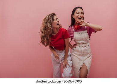Young caucasian lady laughs hard with her pretty asian girlfriend against pink background. Image of 20s girls pretending dancing to music and having fun with each other in studio.