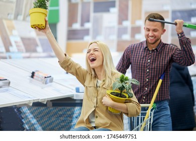 Young Caucasian Lady Sit On Trolley In The Market, Have Fun. Her Husband Roll The Trolley, Woman Hold Plants In Pots, Enjoy Making Purchase