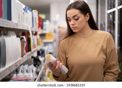 Young Caucasian Lady With A Trolley And Long Hair In A Supermarket Chooses Shampoo. A Woman Buys Groceries And Basic Goods At A Local Organic Store. Copy Space.