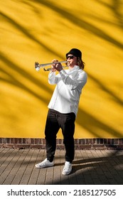 Young Caucasian Long-haired Male Musician In White Shirt, Hat, Sunglasses And Sneakers Playing Classic Jazz On Silver Trumpet With Pleasure Standing Near Yellow Wall On Street Outside On Sunny Day