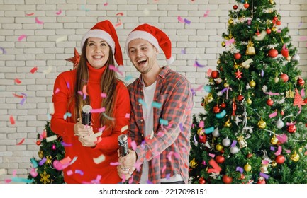 Young Caucasian Lover Couple Husband And Wife In Red Santa Claus Hat And Sweater Standing Laughing Holding Shooting Paper Cracker Confetti Together In Decorated Living Room With Christmas Eve Tree.