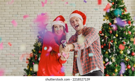 Young Caucasian Lover Couple Husband And Wife In Red Santa Claus Hat And Sweater Standing Laughing Holding Shooting Paper Cracker Confetti Together In Decorated Living Room With Christmas Eve Tree.