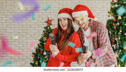 Young Caucasian Lover Couple Husband And Wife In Red Santa Claus Hat And Sweater Standing Laughing Holding Shooting Paper Cracker Confetti Together In Decorated Living Room With Christmas Eve Tree.