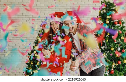 Young Caucasian Lover Couple Husband And Wife In Red Santa Claus Hat And Sweater Standing Laughing Holding Shooting Paper Cracker Confetti Together In Decorated Living Room With Christmas Eve Tree.