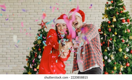 Young Caucasian Lover Couple Husband And Wife In Red Santa Claus Hat And Sweater Standing Laughing Holding Shooting Paper Cracker Confetti Together In Decorated Living Room With Christmas Eve Tree.