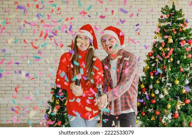 Young Caucasian Lover Couple Husband And Wife In Red Santa Claus Hat And Sweater Standing Laughing Holding Shooting Paper Cracker Confetti Together In Decorated Living Room With Christmas Eve Tree.