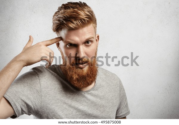 金髪の髭を生やした白人の若い男性で 髪の毛の指で指さす指で 自分のお寺をオセントの意味で指さすのか 頭を使って オセント 灰色のカジュアルtシャツを着たハンサムな男は驚き 少しイライラする の写真素材 今すぐ編集