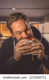 Young Caucasian Male With A Beard Holding A Fresh Hamburger And Taking A Big Bite In A Fast Food Restaurant Environment. Concept Of Unhealthy Snack In The City.