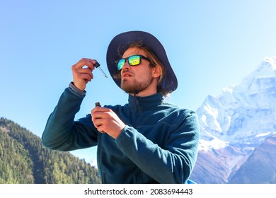 Young caucasian male with a beard wearing a fleece, a hat and sunglasses taking CBD oil from a dripper high up in the Himalaya mountains. Sports enhancement supplement for trekking in high altitude.