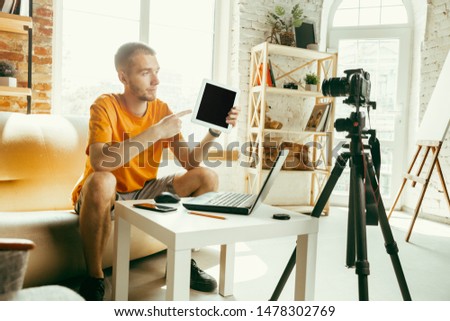 Young caucasian male blogger with professional camera recording video review of tablet at home. Blogging, videoblog, vlogging. Man making vlog or live stream about photo or technical novelty.