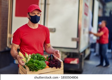 Young Caucasian Male Courier Wearing Black Face Protective Mask Holding Grocery Box, Standing On The Street With Truck On The Background. Food Delivery During Covid 19 Outbreak Concept