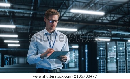 Young Caucasian Male Employee Uses Tablet Computer in System Control Monitoring Center at Night. Evening Office In the Background. Cyber Security and Network Protection Concept