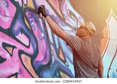 Young caucasian male graffiti artist drawing big street art painting in blue and pink tones. Stylish man in denim shorts and grey t shirt made new graffitti artwork outdoors in bright sunny day - Powered by Shutterstock