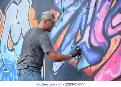 Young caucasian male graffiti artist drawing big street art painting in blue and pink tones. Stylish man in denim shorts and grey t shirt made new graffitti artwork outdoors in bright sunny day - Powered by Shutterstock