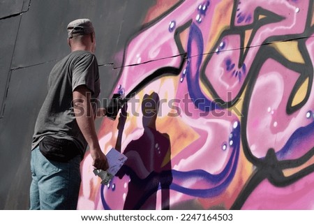 Young caucasian male graffiti artist drawing big street art painting in blue and pink tones. Stylish man in denim shorts and grey t shirt made new graffitti artwork outdoors in bright sunny day