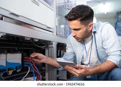Young Caucasian male industrial engineer in blue shirt squatting touch and checking wire cables that is plugged on the back of the machine with the digital tablet in his hand in an industrial factory.