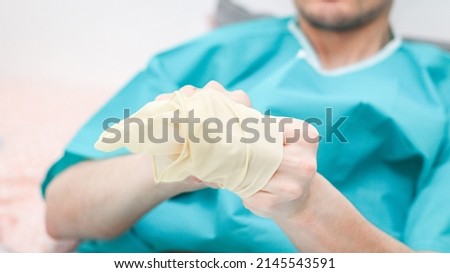 Young caucasian male patient in medical disposable pajamas puts sterile surgical white gloves on his hands to replace the coloplast after surgery at home,  side view. Concept of medicine, abdominal di