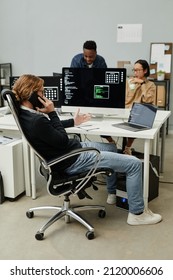 Young Caucasian male programmer calling by mobile phone by workplace while his two colleagues discussing data on computer screen - Shutterstock ID 2120006606