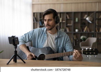 Young Caucasian male singer in headphones hold play guitar record new single on smartphone at home studio. Millennial man artist use musical instrument sing shoot music video on cellphone camera.