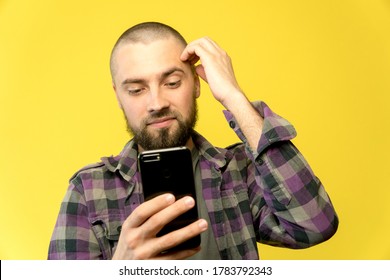 A Young Caucasian Man With A Beard And A Bald Head Holding A Phone In His Hands On A Yellow Background. Guy Holds His Head By Head, Smiles, And Looking On Screen. Smart Concept With Copy Space.