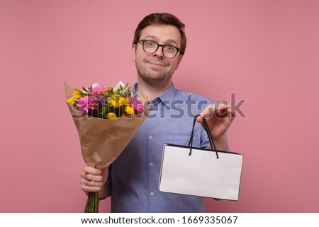 Young caucasian man in blue shirt holding flowers and package as gift for his mother or girlfriend on birthday. Anniversary present concept. Studio shot on colored wall.