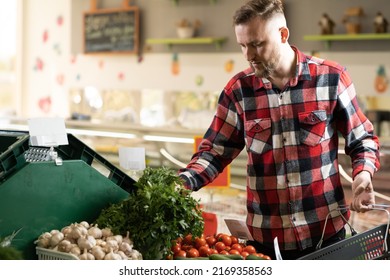 Young Caucasian Man Buying Vegetables In Grocery Section At Supermarket. Choose Vegetables And Greens While Holding Grocery Basket. Millennial Man Shopping Veggies At Supermarket.