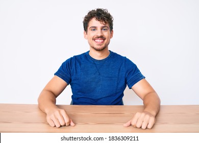 Young Caucasian Man With Curly Hair Wearing Casual Clothes Sitting On The Table Looking Positive And Happy Standing And Smiling With A Confident Smile Showing Teeth 