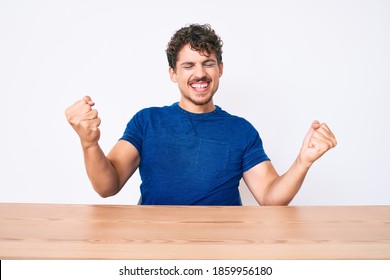 Young Caucasian Man With Curly Hair Wearing Casual Clothes Sitting On The Table Very Happy And Excited Doing Winner Gesture With Arms Raised, Smiling And Screaming For Success. Celebration Concept. 