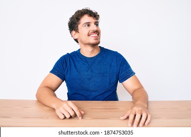 Young Caucasian Man With Curly Hair Wearing Casual Clothes Sitting On The Table Looking Away To Side With Smile On Face, Natural Expression. Laughing Confident. 