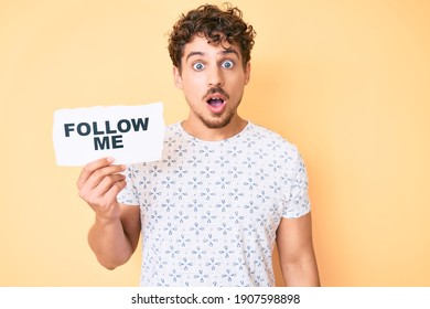 Young caucasian man with curly hair holding follow me message paper scared and amazed with open mouth for surprise, disbelief face 