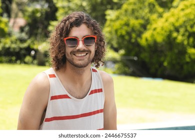 A young Caucasian man with curly hair smiles outdoors with copy space at home. He wears orange-framed sunglasses, striped tank top, and exudes a relaxed vibe, unaltered. - Powered by Shutterstock