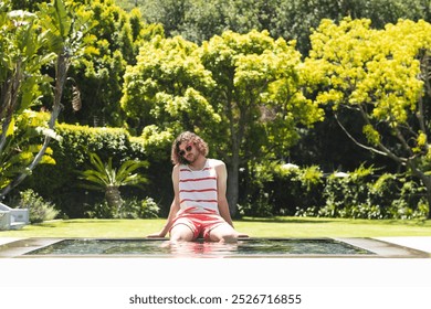 Young Caucasian man with curly hair sits by a pool, wearing a striped tank top and sunglasses, with copy space. Surrounded by lush greenery, he enjoys a sunny day in a tranquil garden setting. - Powered by Shutterstock