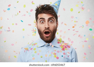 Young Caucasian Man Dressed In Formal Shirt And Having Party Cap On Head, Celebrating His Birtday, Looking With Bugged Eyes And Widely Opened Mouth Into Camera Being Surprised To Recieve Many Presents