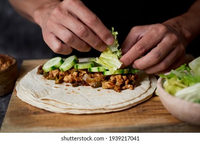 a young caucasian man fills a durum or a burrito, with chicken meat cooked with onion and red and green pepper, and fresh lettuce and some slices of raw cucumber