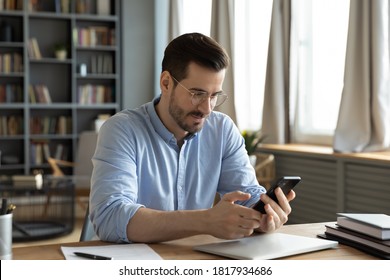 Young Caucasian Man In Glasses Sit At Desk At Home Office Look At Smartphone Screen Consult Client Online. Millennial Male Use Modern Cellphone Gadget, Texting Or Messaging On Wireless Internet.