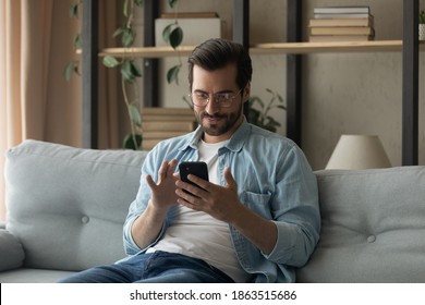 Young Caucasian Man In Glasses Sit Rest On Sofa At Home Look At Cellphone Screen Texting Or Messaging. Millennial Male Relax On Couch In Living Room Browse Wireless Internet On Smartphone Gadget.