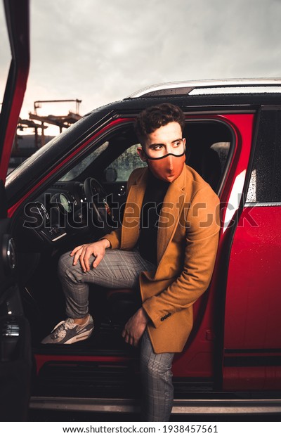 Young caucasian man going out from a red car wearing\
a mask on a parking lot.