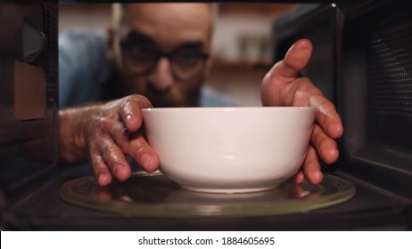 Young Caucasian Man Heating Food In Microwave Oven. View From Inside Microwave Of Bearded Guy Opening Door And Taking Warm Bowl With Meal For Dinner